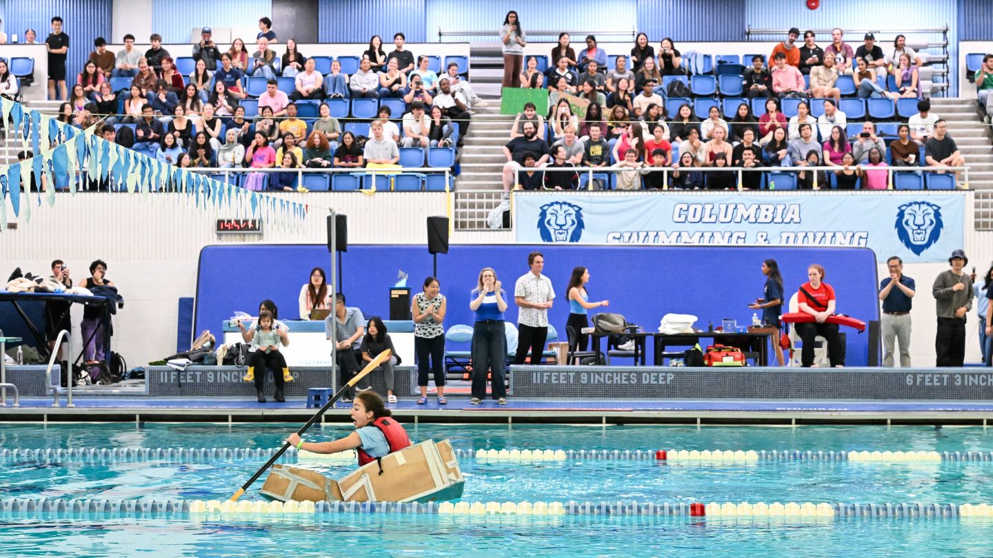 Columbia Engineering student paddles a cardboard boat across Uris Pool as spectators watch from the stands.