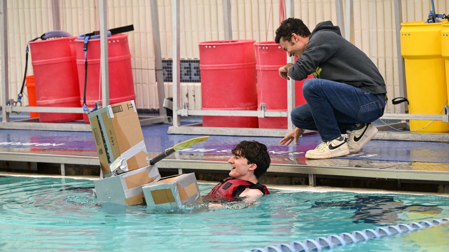 Student smiles in Uris Pool beside a partially submerged cardboard boat as another student laughs nearby from the pool deck.