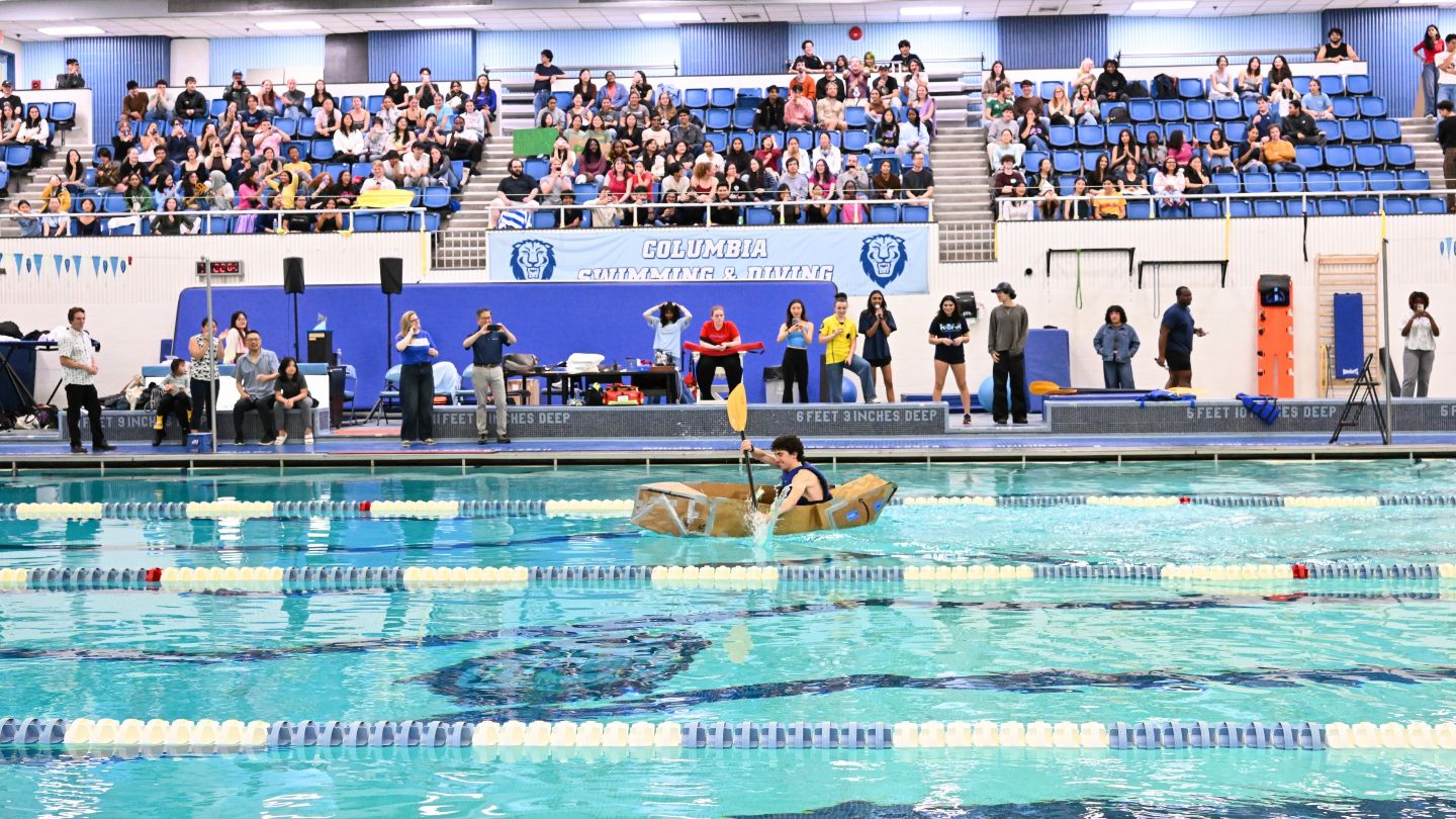 Columbia Engineering student paddles a cardboard boat across Uris Pool as spectators watch from the stands and pool deck.