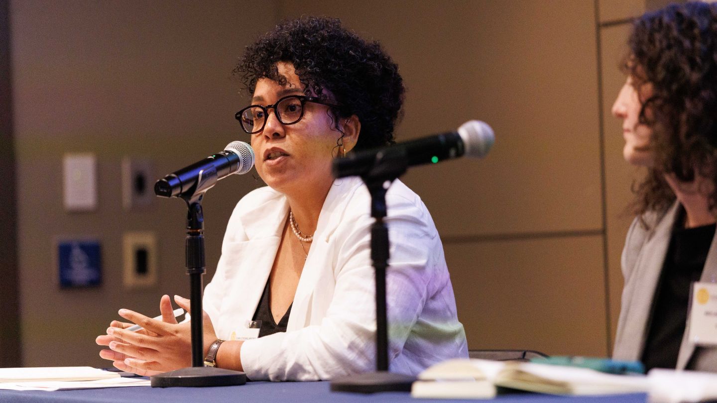 Juliana Carneiro speaking into a microphone while seated at a panel table, with another panelist beside her.