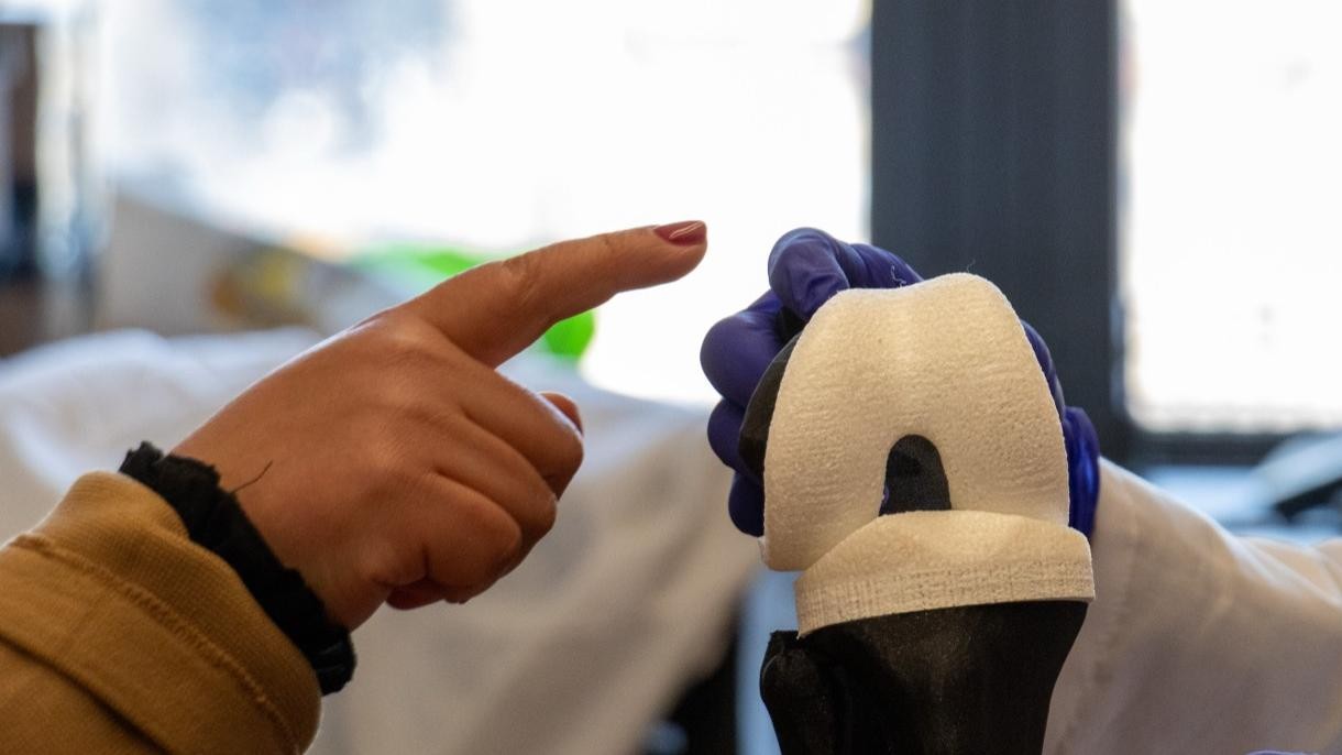 Gloved researcher holds a NovaKnee prototype while another person points toward it in a lab.