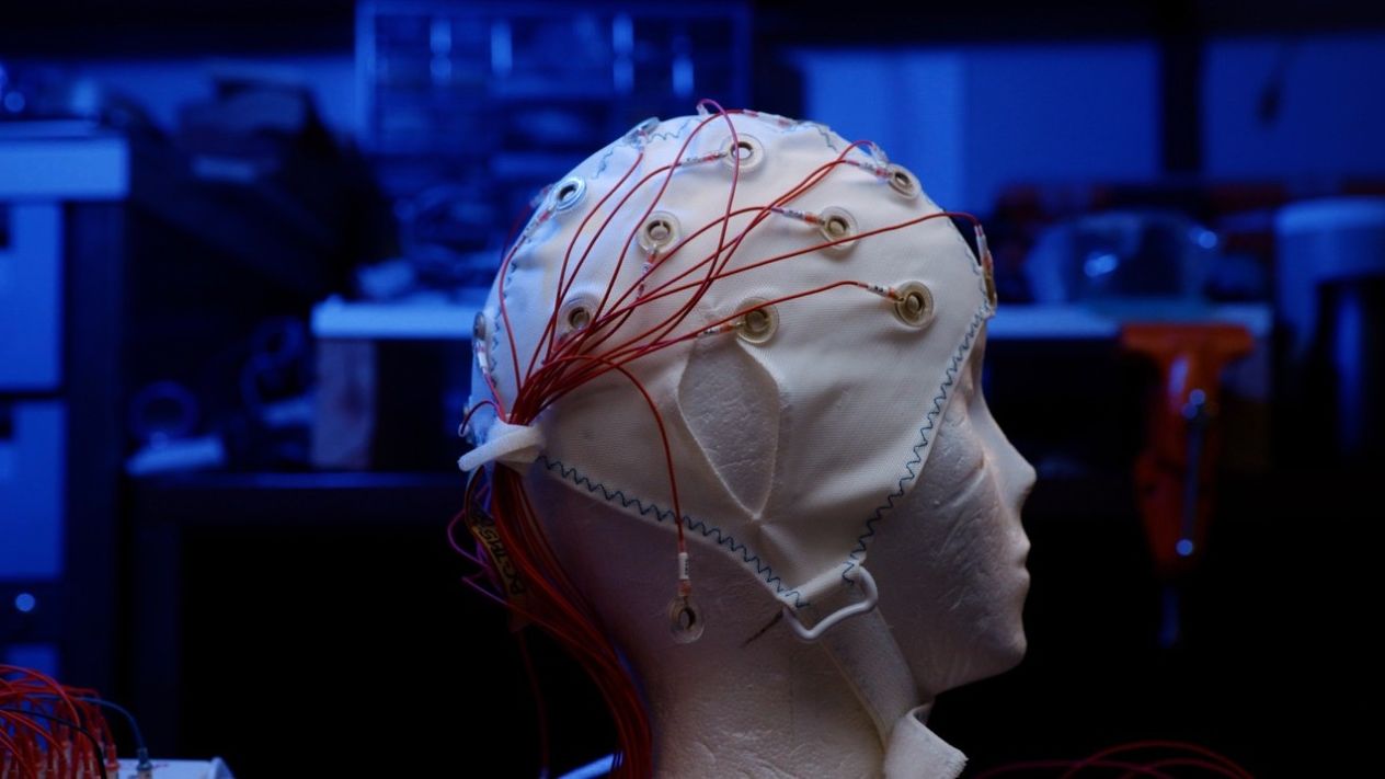 White electrode cap with red wires fitted over a model of a human head in a dark laboratory.