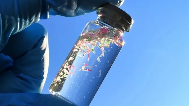 Gloved hand holds a vial of water containing colorful microplastic particles.