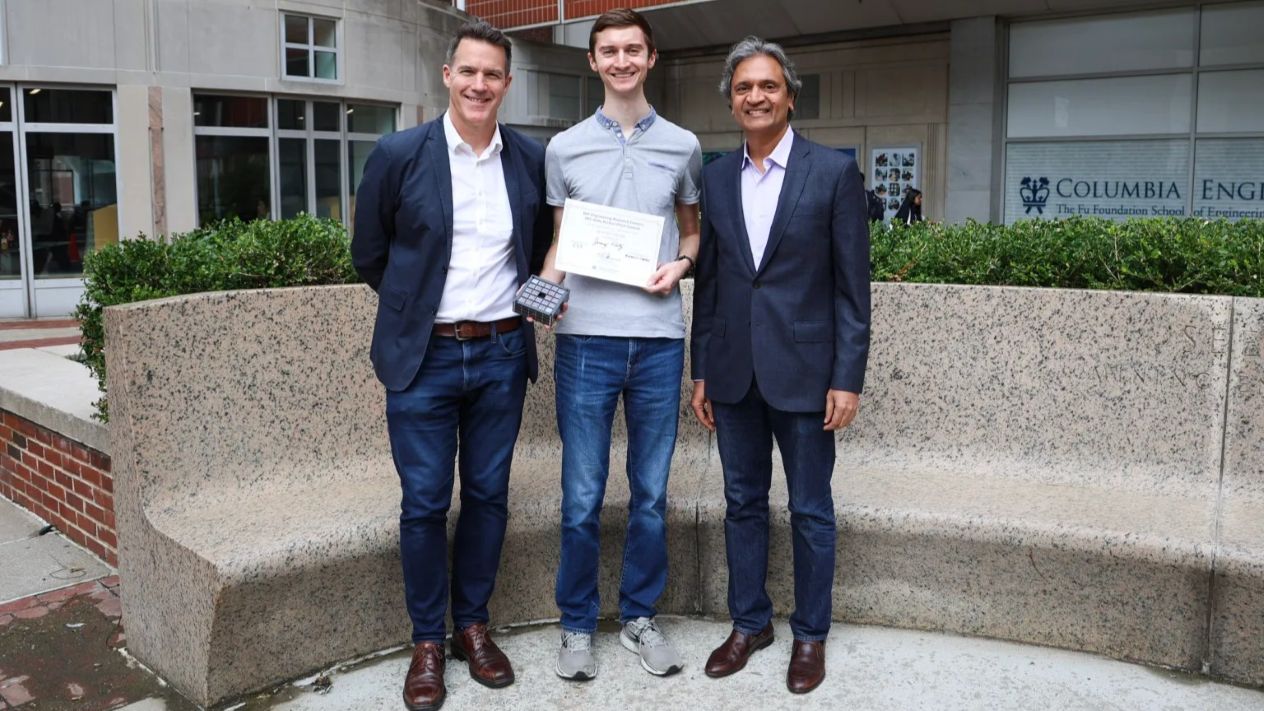CS3 Director Andrew Smyth, Jeremy Klotz, and Prof. Shree Nayar stand together outside Mudd at Columbia Engineering, with Klotz holding a minimalist camera prototype and his second-place certificate.