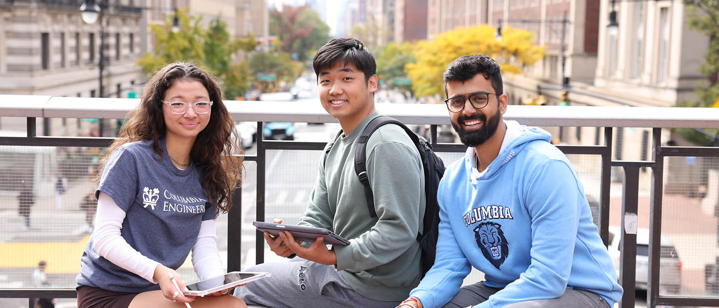 Three students smile while sitting on a bridge overlooking a busy New York City street.