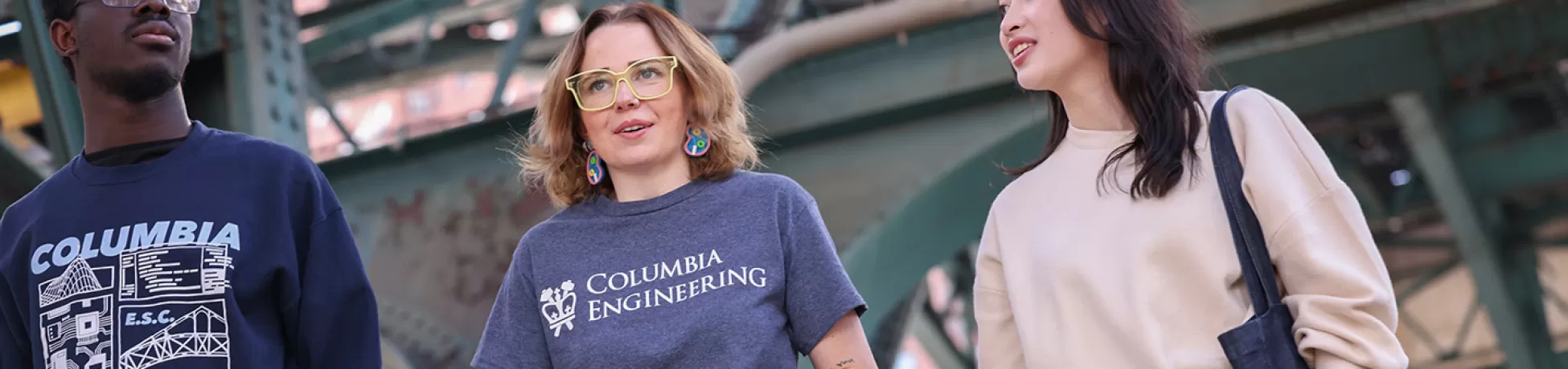 Three Columbia Engineering students walk together under a large bridge in New York City.