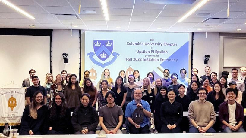 Three rows of people smiling for a photo. In the background, a large screen reads "Columbia University Chapter of Upsilon Pi Epsilon Fall 2023 Initiation Ceremony