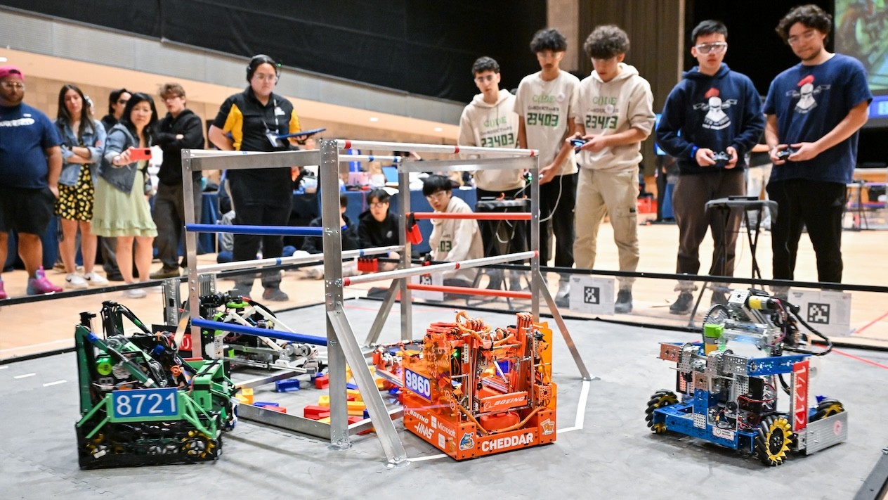 Four robots in a boxing ring-like space. Out of focus, six students hold remote controls.