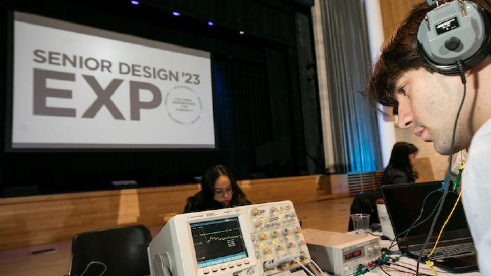 A student wearing headphones, a sign reads "Senior Design '23 Expo"