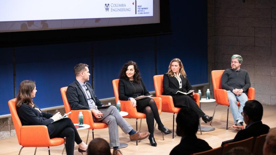 Five panelists sitting in a row on-stage. The backs of audience members' heads appear in the foreground.