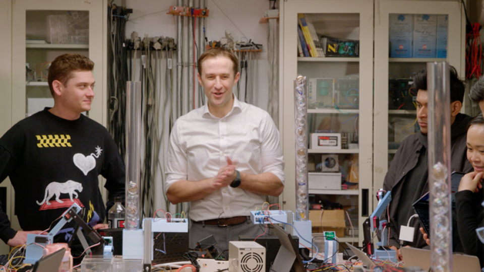 A professor with their hands together standing in a room cluttered with wires and electronics. Students look at them from either side.