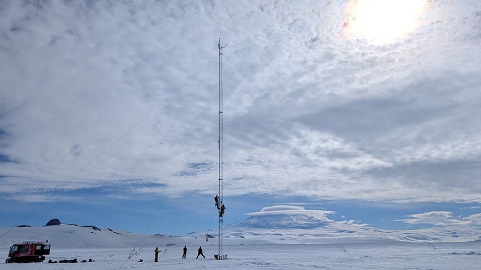 Two people climb a radio tower in a snowy, antarctic landscape