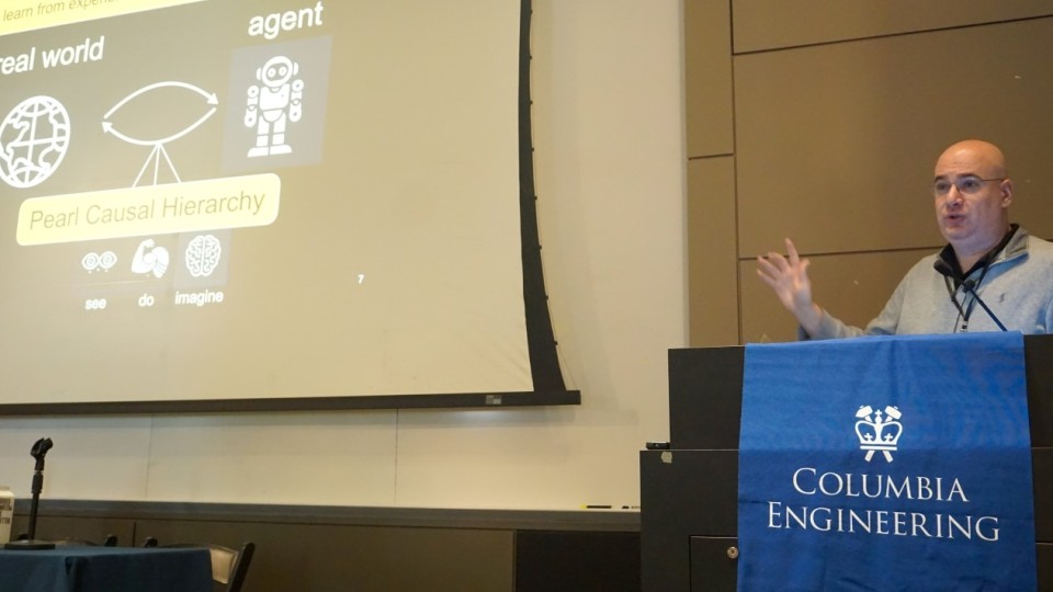 Elias Bareinboim speaking into a microphone at a podium with a Columbia Engineering banner. The screen to his right reads "Pearl Causal Hierarchy"