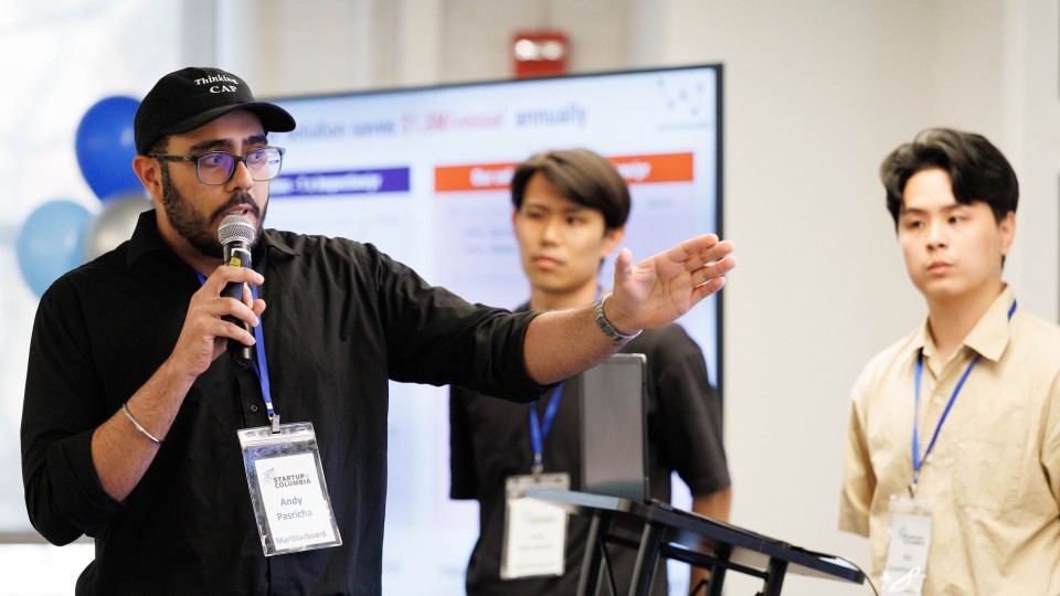  man wearing a black cap and glasses speaks into a handheld microphone and gestures with his arm extended during a presentation; two other participants with name badges stand nearby, with a large screen and blue-and-white balloons in the background.