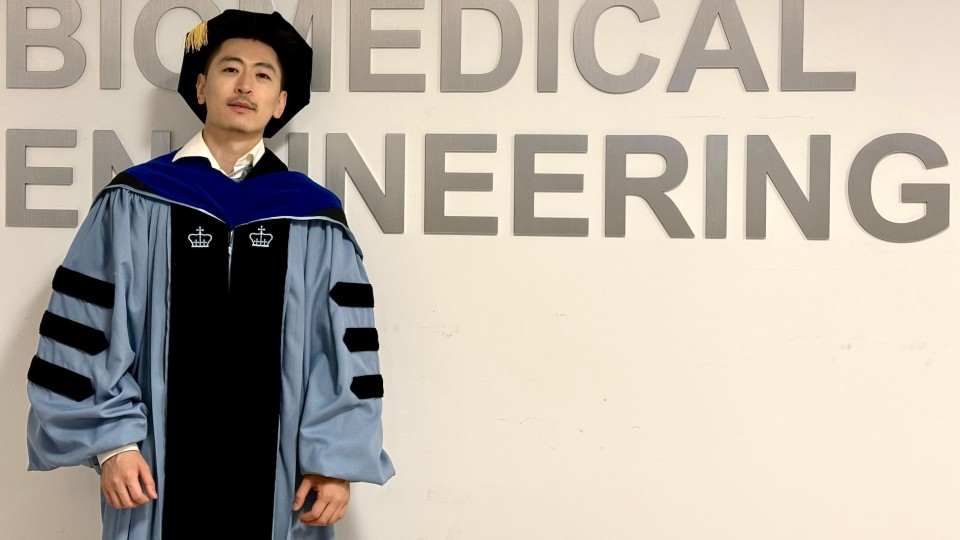 A man wearing Columbia University PhD graduation regalia posing for a photo in front of a sign that reads "Biomedical Engineering"