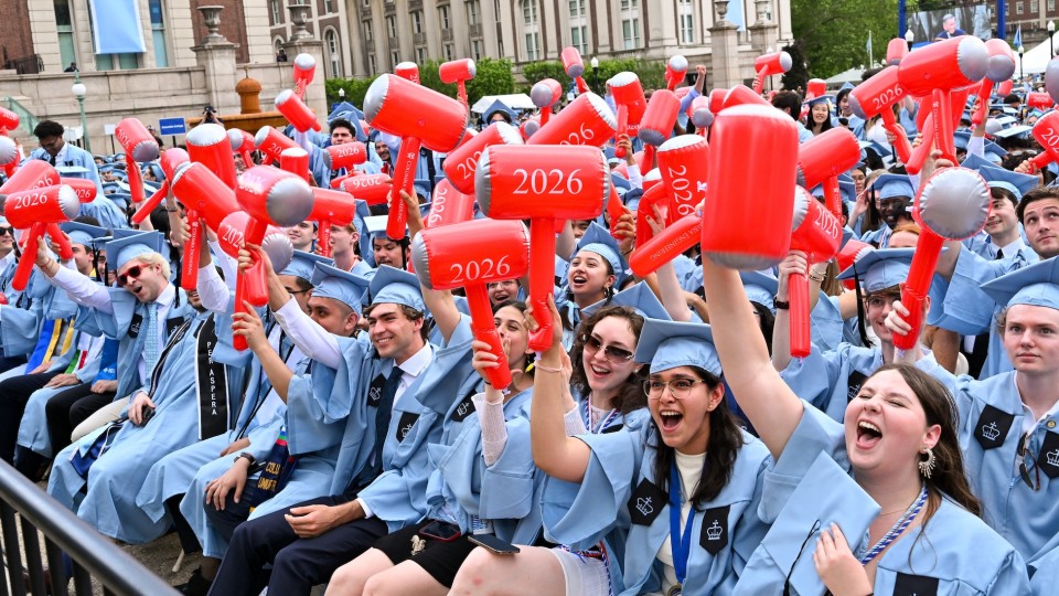 A large crowd of Columbia Engineering graduates in light blue caps and gowns cheer and wave red inflatable mallets printed with “2026” during an outdoor graduation ceremony