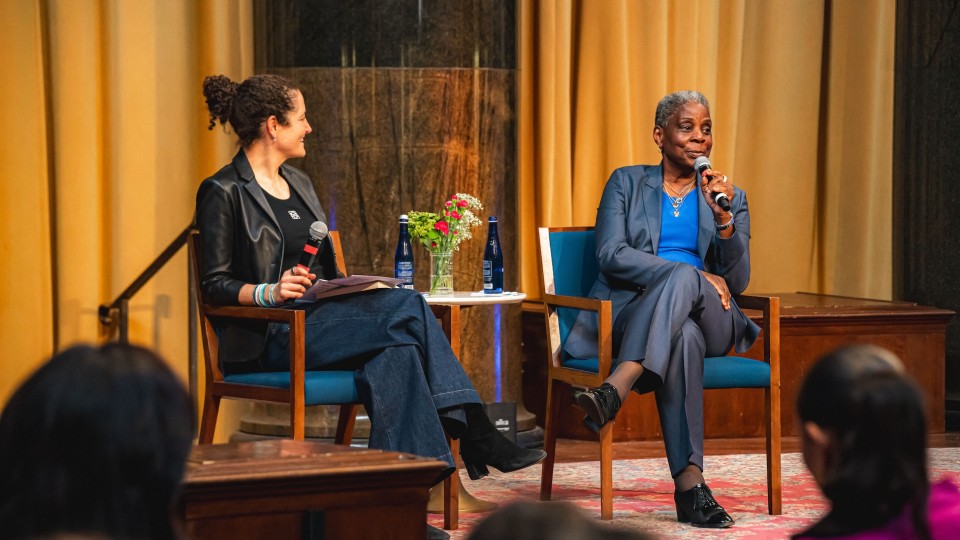 Two women sit on stage in armchairs during a public talk; one listens with a microphone in hand while the other, in a blue suit, speaks into a microphone. A small table between them holds water bottles and a vase of flowers, with an audience visible in the foreground.