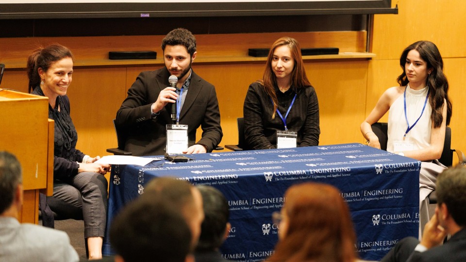 Four panelists sit at a table with a “Columbia Engineering” tablecloth during a talk; one man in a suit speaks into a handheld microphone while three women listen, with an audience visible in the foreground.