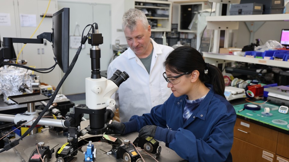 n a busy laboratory, a scientist in a blue lab coat and safety glasses works at a microscope-and-monitor workstation, adjusting small components on an optical table while a colleague in a white lab coat stands beside them observing.