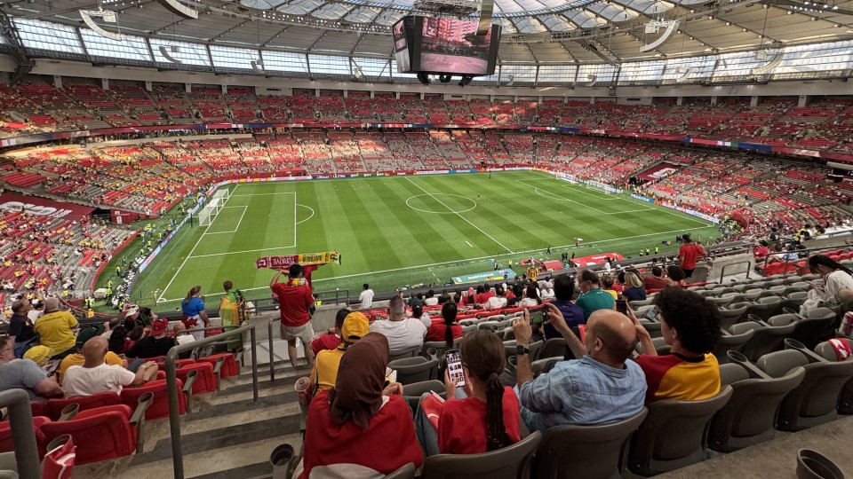 Wide view from the stands inside a large covered stadium before a soccer match. A green pitch with white lines sits below, surrounded by mostly red seats with scattered fans; a big video screen hangs from the roof. In the foreground, spectators in red and yellow jerseys sit and take photos, and one fan holds a scarf up toward the field.