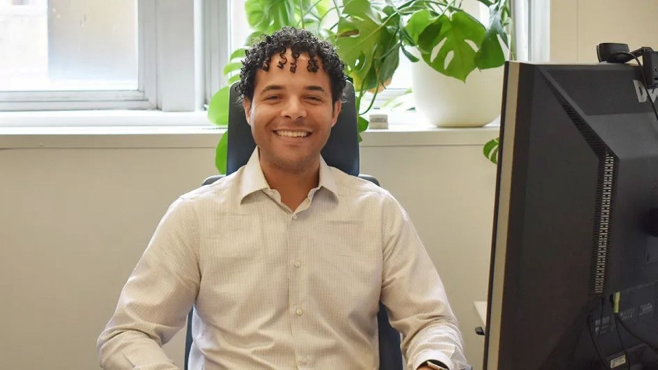 Brian A. Smith smiling in his office, seated near a window with natural light, a potted plant and a computer monitor visible on his desk