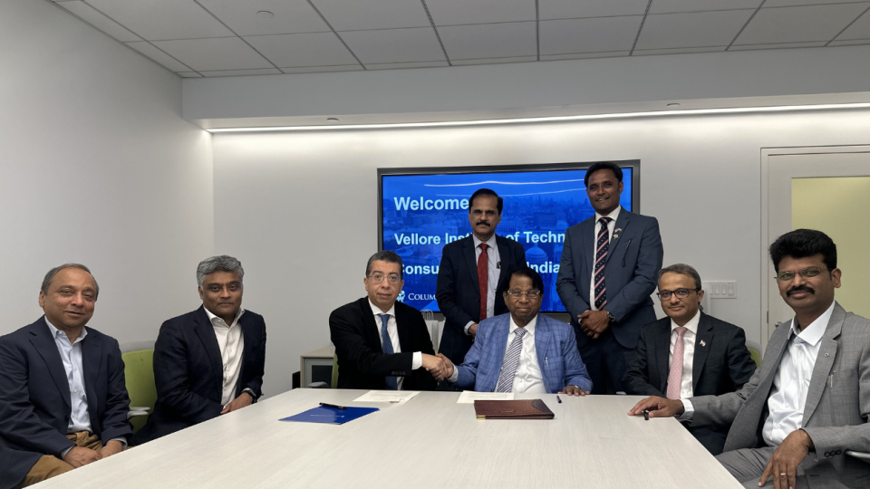 Soulaymane Kachani shakes hands with a Vellore Institute of Technology official after signing a Memorandum of Understanding, with Hardeep Johar and other representatives from Columbia Engineering, VIT, and the VIT Alumni Association of North America seated around the table, and a welcome screen for the VIT delegation visible behind them
