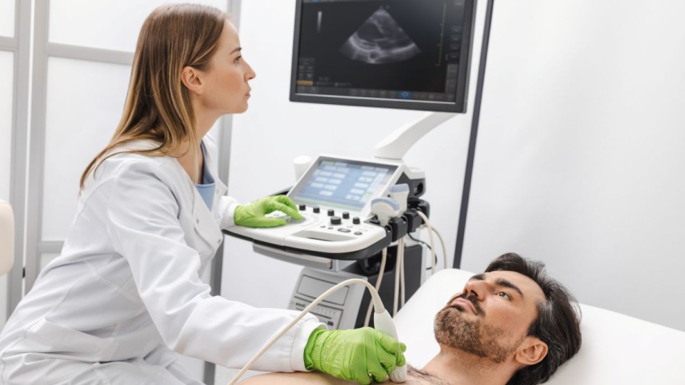 A female healthcare provider in a white coat and green gloves performs a heart ultrasound on a male patient lying on an exam table, with an echocardiogram image displayed on the monitor screen.