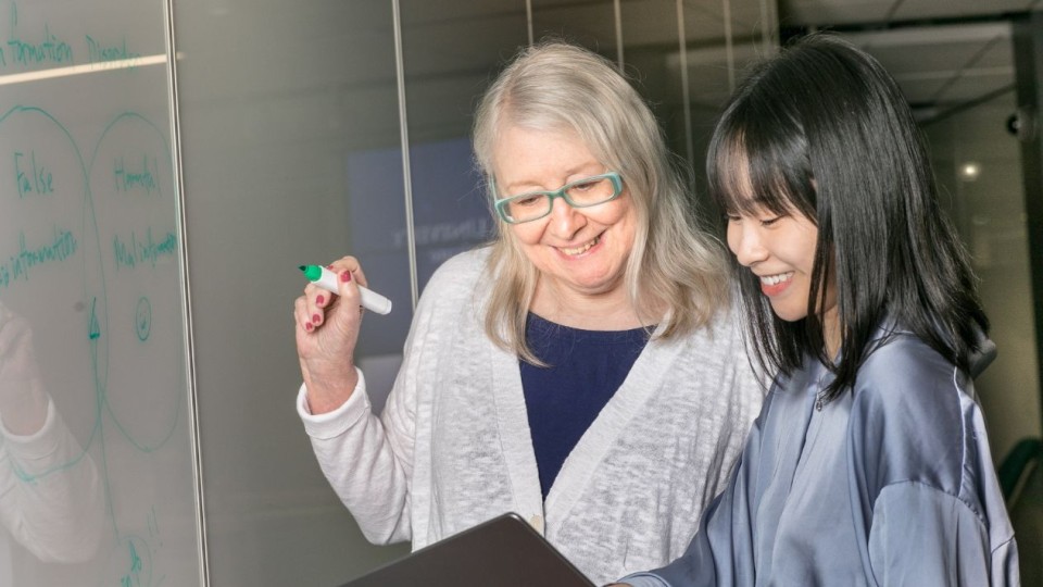 Julia Hirschberg smiling and looking down at a Mac laptop with one of her students while holding a green marker in a classroom setting