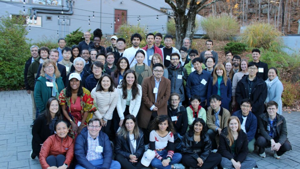 Large group of attendees, some wearing name badges, posed in rows outdoors in front of a building with autumn foliage in the background.