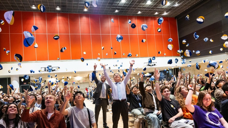 Dean Shih-Fu Chang, Barclay Morrison III, and hundreds of students throw beanies into the air in an auditorium