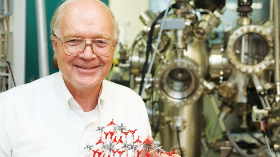 Richard M. Osgood holding a red and silver molecular model in front of laboratory equipment.