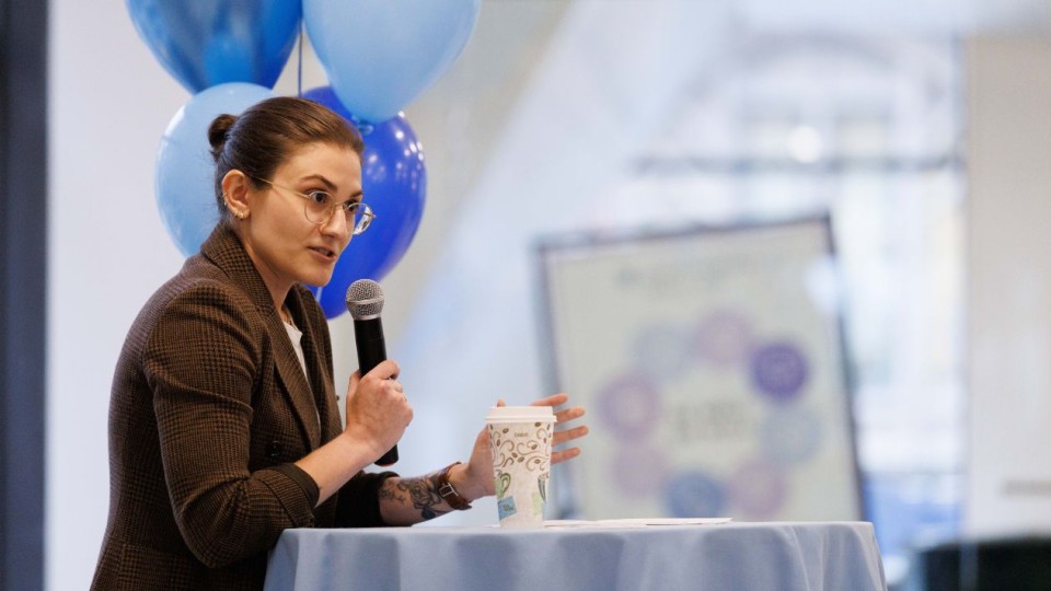 Angela McNeal speaks into a microphone while seated at a light blue table, with a coffee cup in front of them and blue balloons behind them.