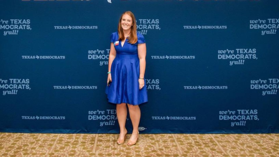 Lauren Pully stands in front of a Texas Democrats backdrop wearing a blue dress.