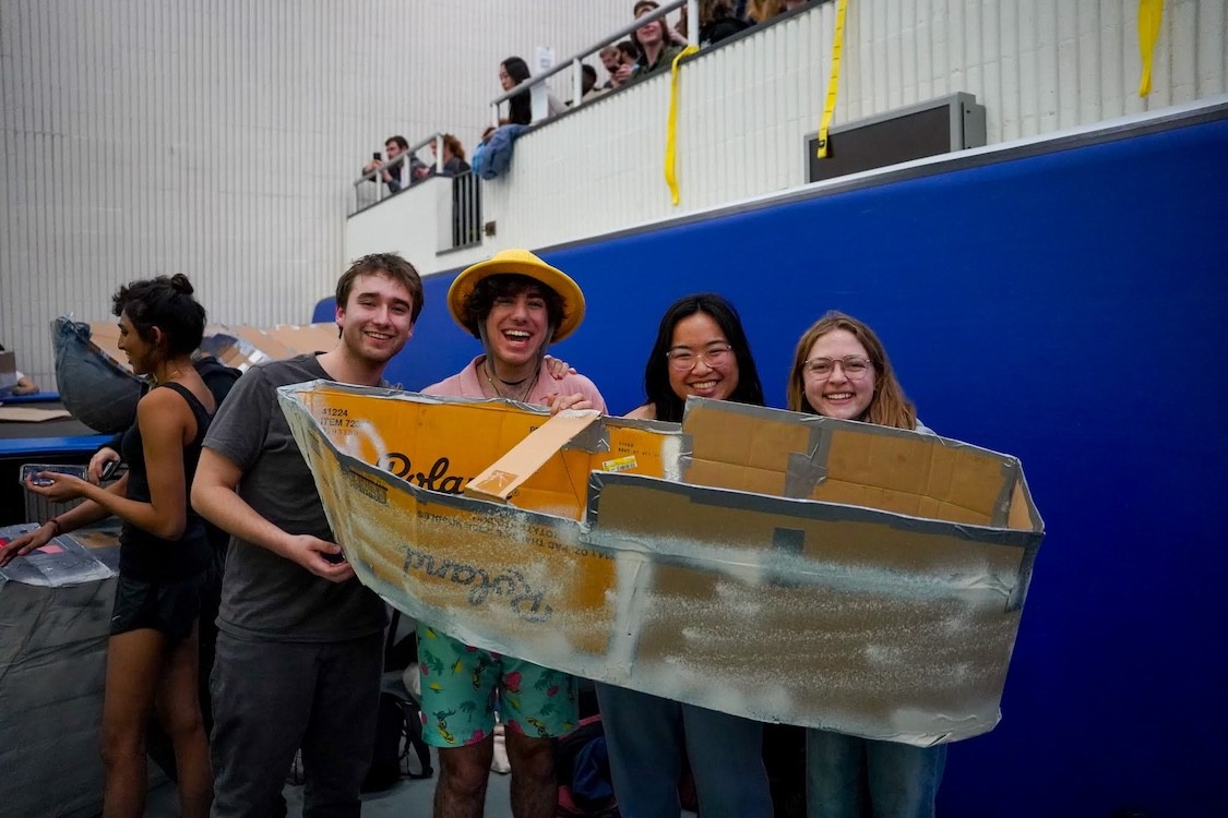Four people smiling while holding a boat made of cardboard and duct tape.