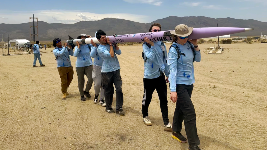 Six people carrying a purple rocket in the desert