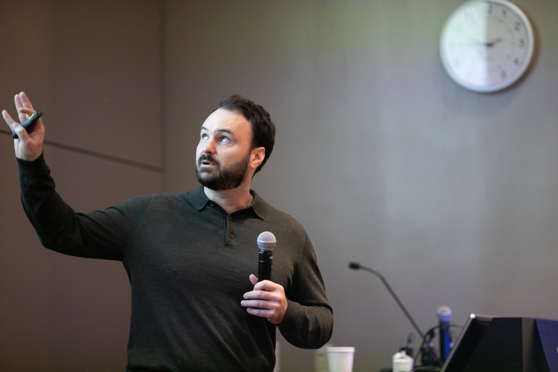 speaker holding a microphone while gesturing to a presentation beside him