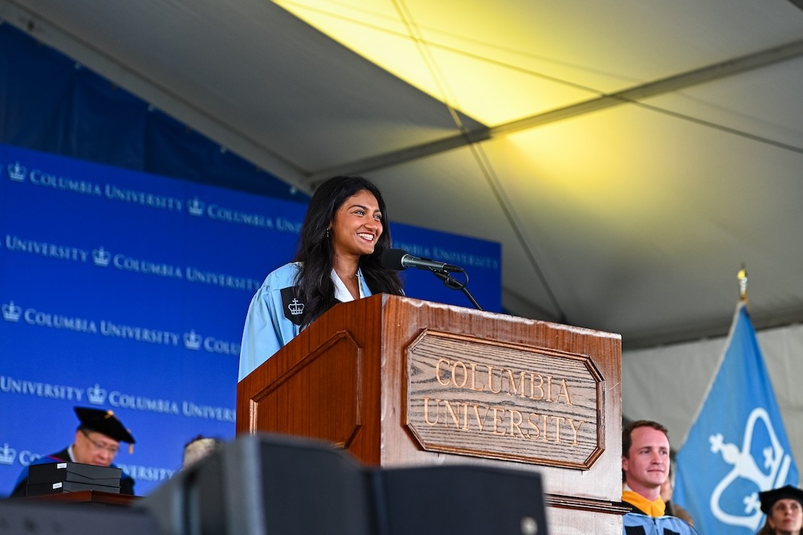 Divyadarshini Thirugnanasambandan smiling in graduation regalia behind a podium that reads "Columbia University" with a microphone.