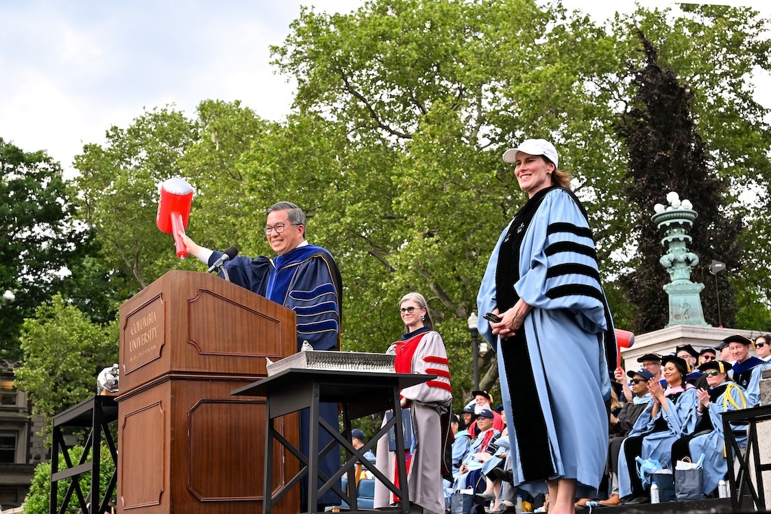Dean Shih-Fu Chang in graduation regalia standing at a podium and holding up an inflatable red mallet as Columbia faculty look on.