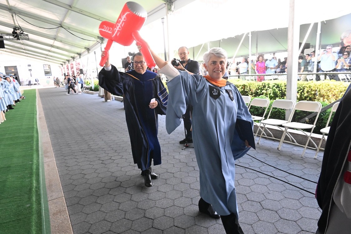 Columbia Engineering Dean Shih-Fu Chang and Undergraduate Class Day speaker Angeliki Frangou in academic robes walk under a large tent at a graduation ceremony, each holding up a red inflatable mallet printed with “2026,” while graduates line the walkway and spectators watch in the background.
