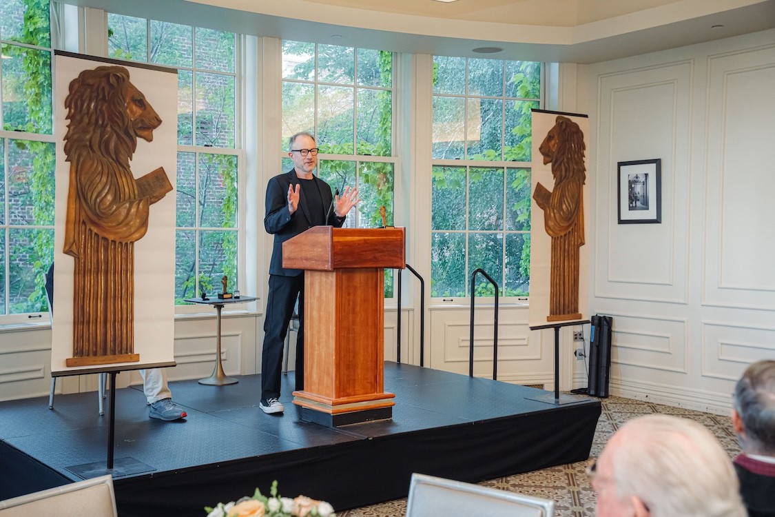Harry West stands behind a podium in a bright room with large windows, speaking between two tall banners featuring carved lion figures during a Columbia Engineering alumni reunion program.