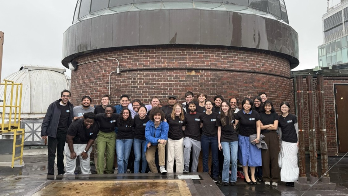 A group of about 20 people pose together on a rainy rooftop in front of a large brick observatory topped with a dark dome. Most are wearing matching black T‑shirts, standing shoulder to shoulder and smiling; wet pavement and nearby rooftop equipment are visible under an overcast sky