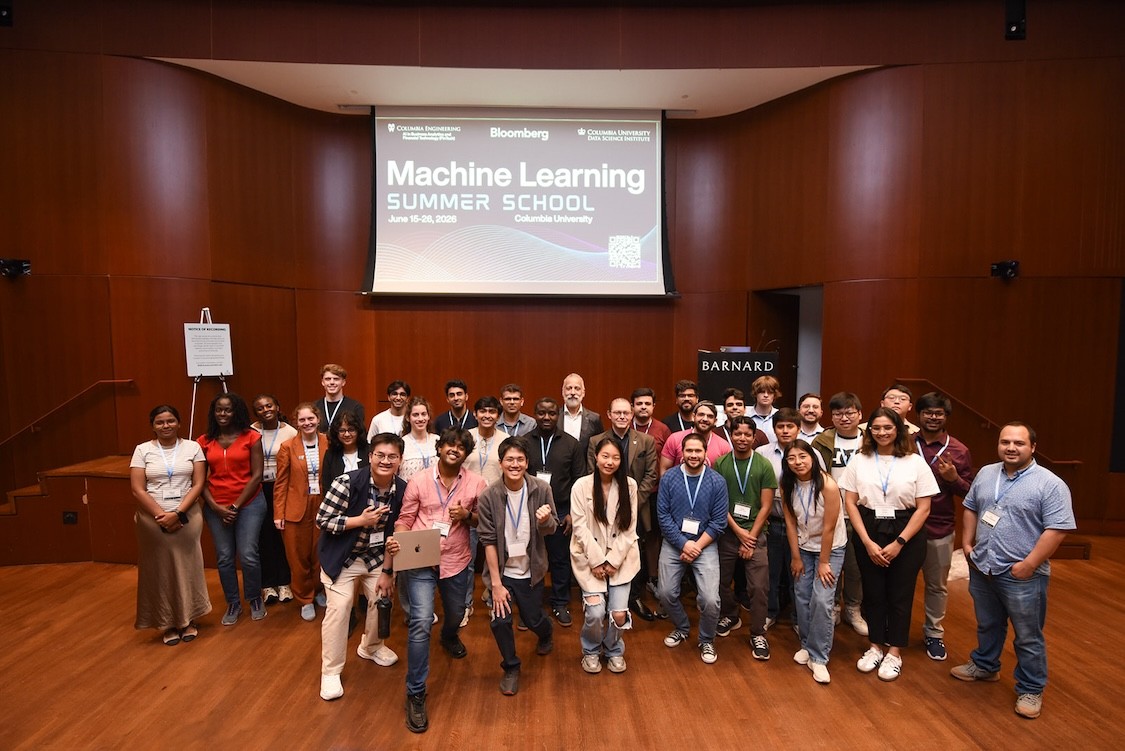 Group photo of about 30–35 people wearing lanyards on a wood-paneled auditorium stage beneath a projected slide reading “Machine Learning Summer School,” with a Barnard podium to the right.