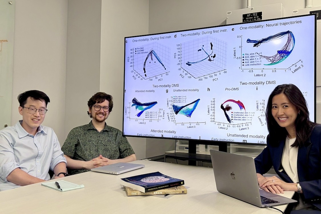 Three people sit at a conference table in an office meeting room, smiling toward the camera. Behind them, a large wall-mounted monitor displays multiple scientific graphs with labels such as “One-modality,” “Two-modality,” and “Neural trajectories.” On the table are a laptop, a notepad and pen, and a small stack of books.