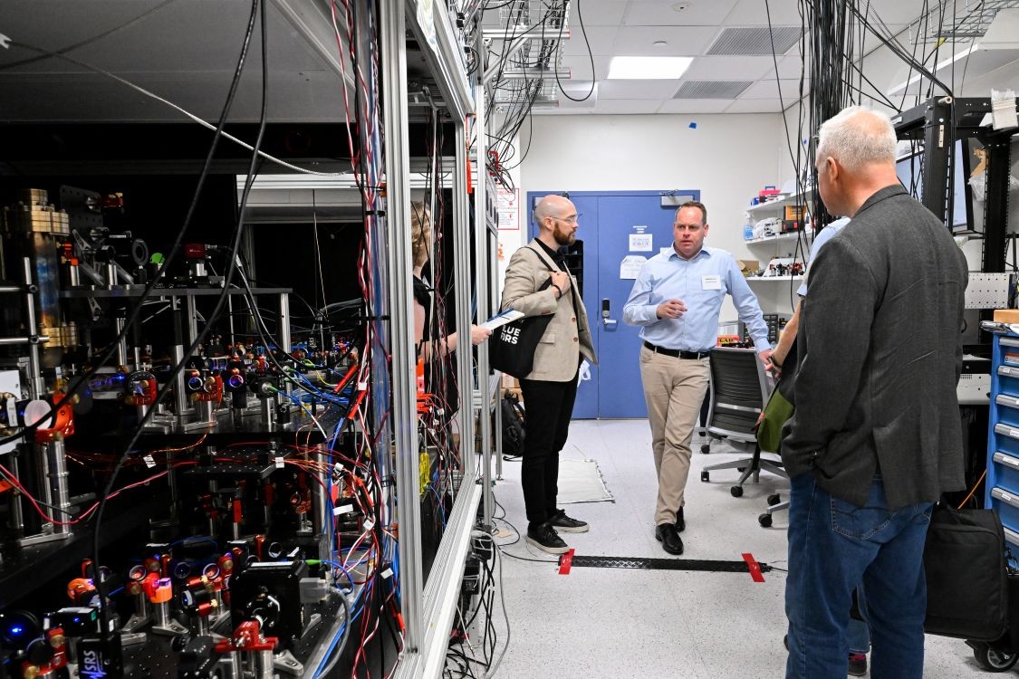 Sebastian Will gestures while explaining his research to two visitors during a lab tour, with laser and optics equipment visible on the other side of the room.
