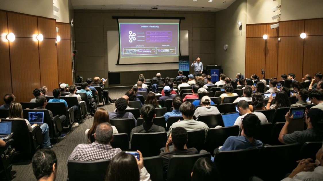 Jun Rao speaking to an audience in Davis Auditorium with a presentation on stream processing displayed on a large screen.