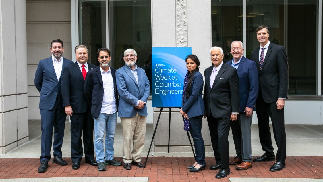 Eight speakers and panel moderators pose together in front of a Climate Week at Columbia Engineering sign outside a glass-fronted building.