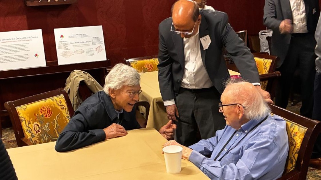 Mischa Schwartz sits at a table talking with Debasis Mitra as Krishan Sabnani stands nearby.