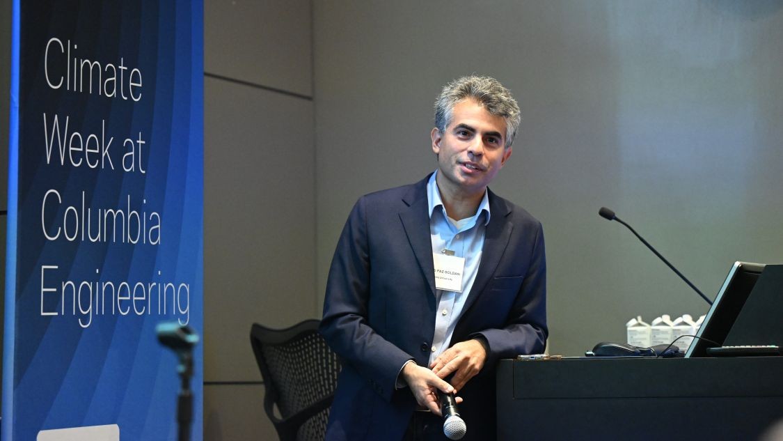 Carlos Paz-Soldan holds a microphone while leaning against a podium beside a Climate Week at Columbia Engineering sign.