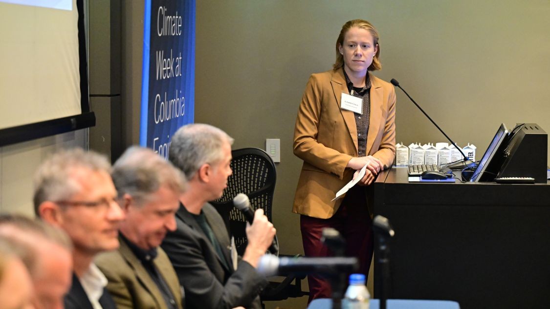 Elizabeth Paul stands beside a podium facing a panel of four people as one panelist speaks into a microphone.