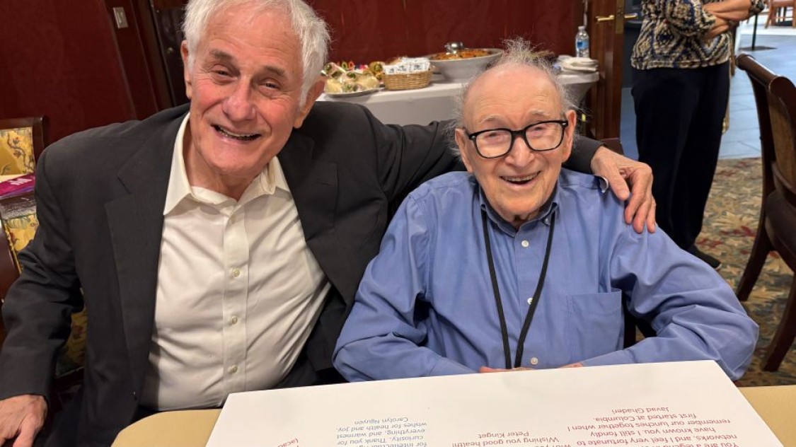 Mischa Schwarz and Zvi Galil smile together at a table, with Zvi’s arm around Mischa.